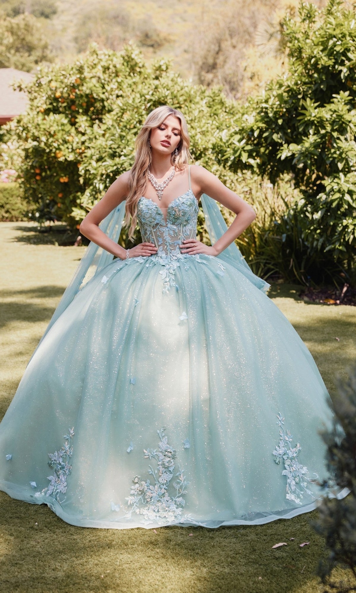 A woman stands outdoors on grass, wearing the Sage Green Quinceanera Dress: Juliet JT1466H, featuring a sheer corset bodice and floral appliqués. Lush green bushes and trees form the backdrop for her elegant ball gown skirt.